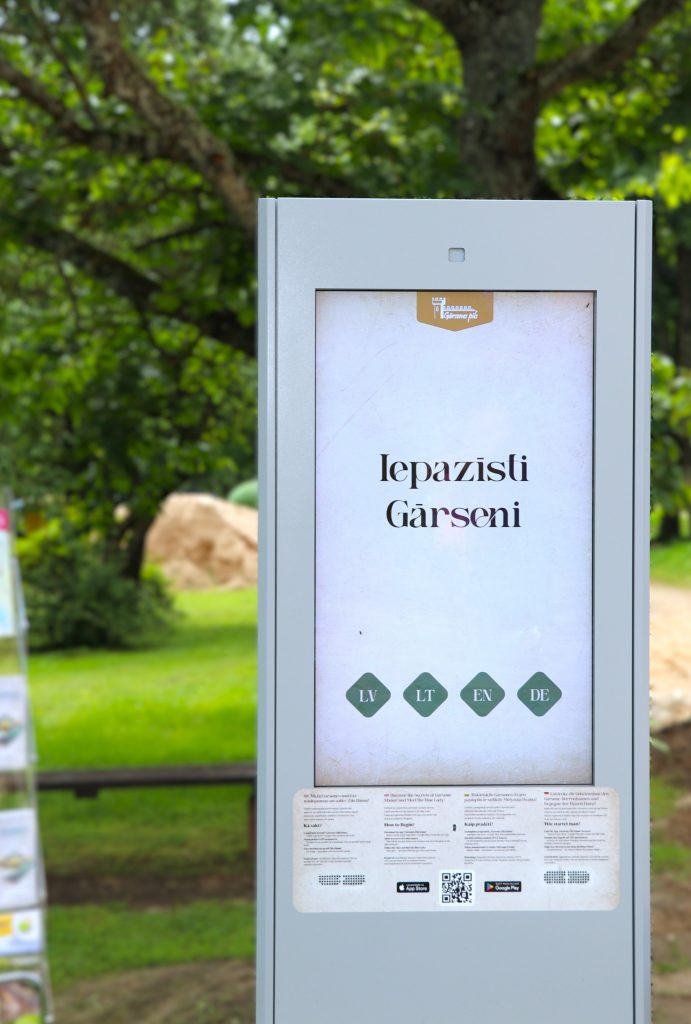 Gārsenes gids viedtālruņa ekrānā muižas parkā